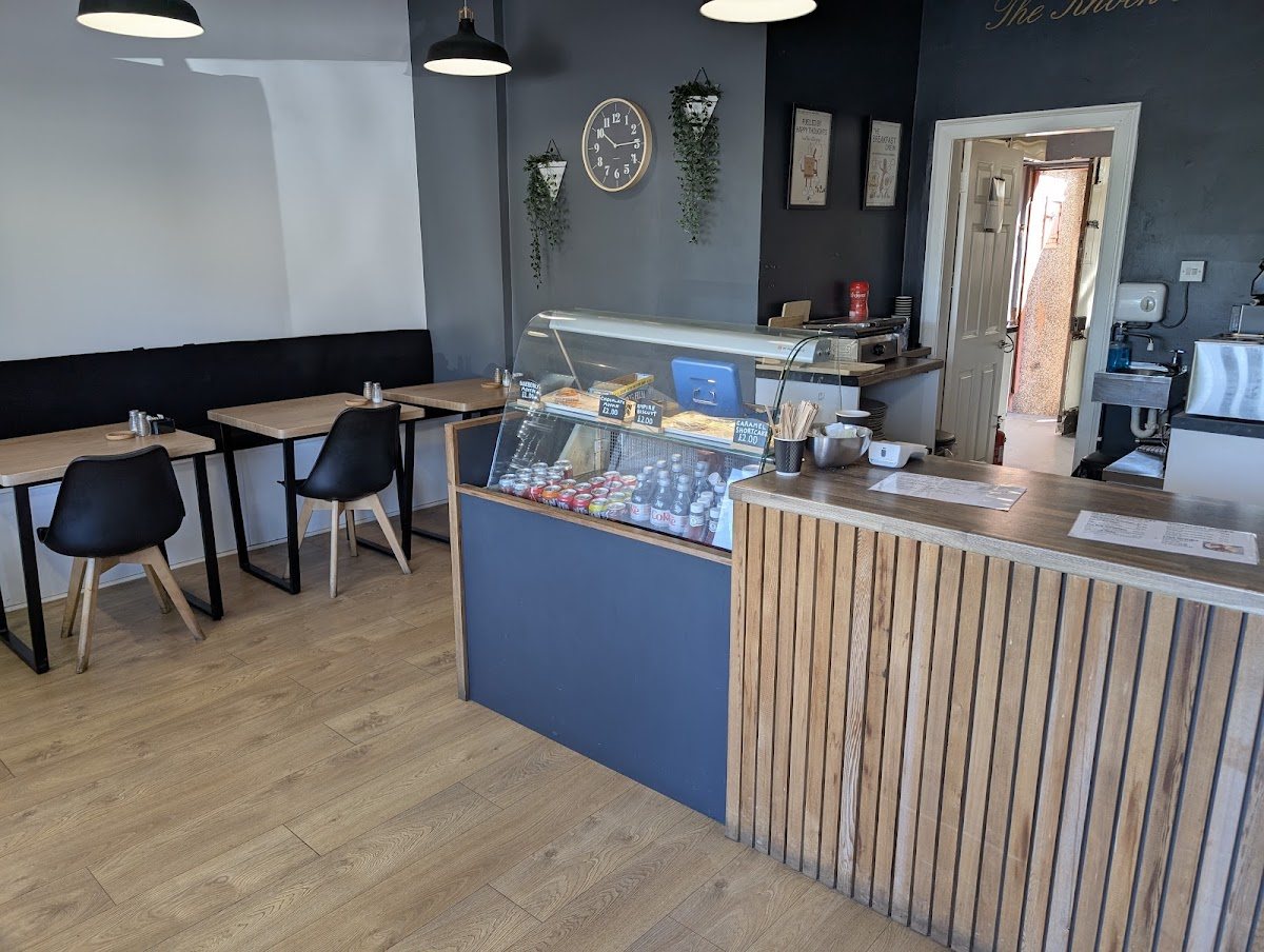 Inside Cafe Frokada: charcoal walls with a round clock, hanging plants and framed prints, a glass counter cabinet, a slatted oak counter, and light wood tables with black chairs.